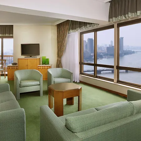 Ramses Hilton Hotel & Casino Cairo Exterior photo The image shows a hotel suite or a well-furnished living area. It features a comfortable seating arrangement with four green chairs and a green sofa. In the center, there is a wooden coffee table. A large window provides a view of a city skyline and 